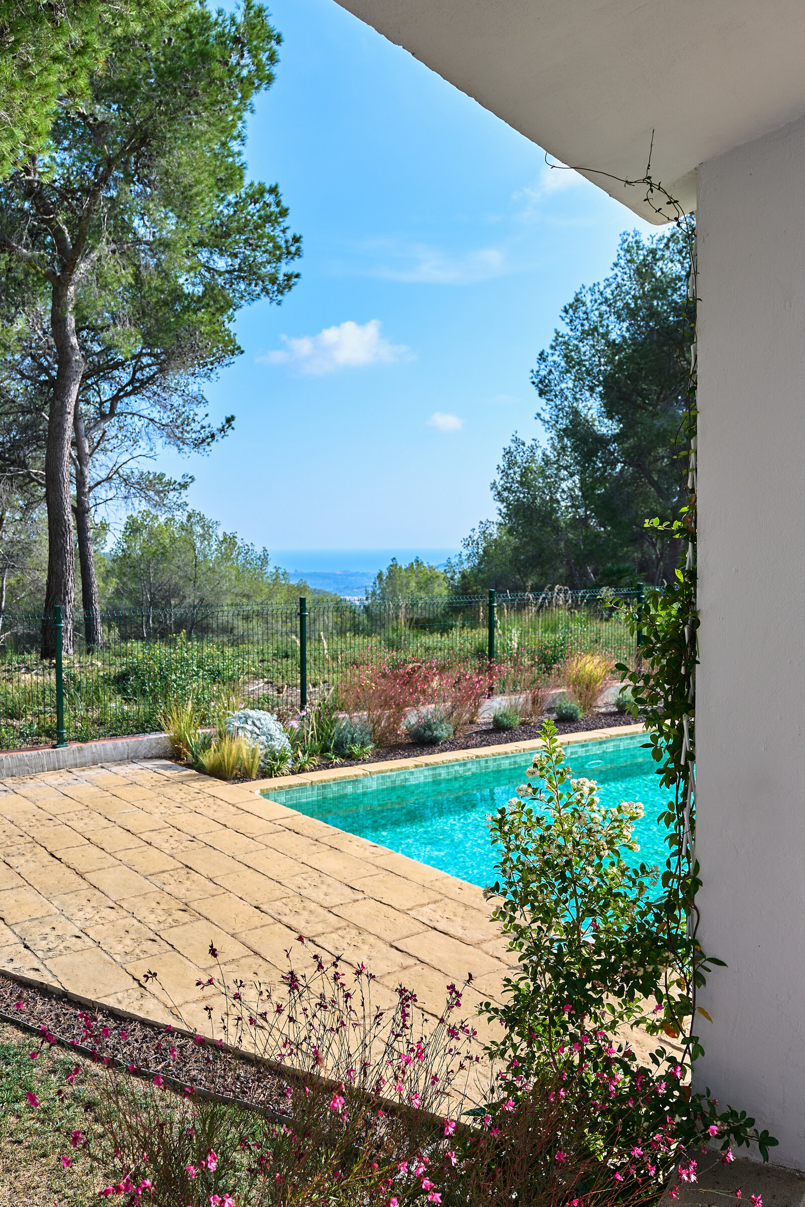 The pool beside a paved terrace, seen from the house, with pines and a distant sea view behind.