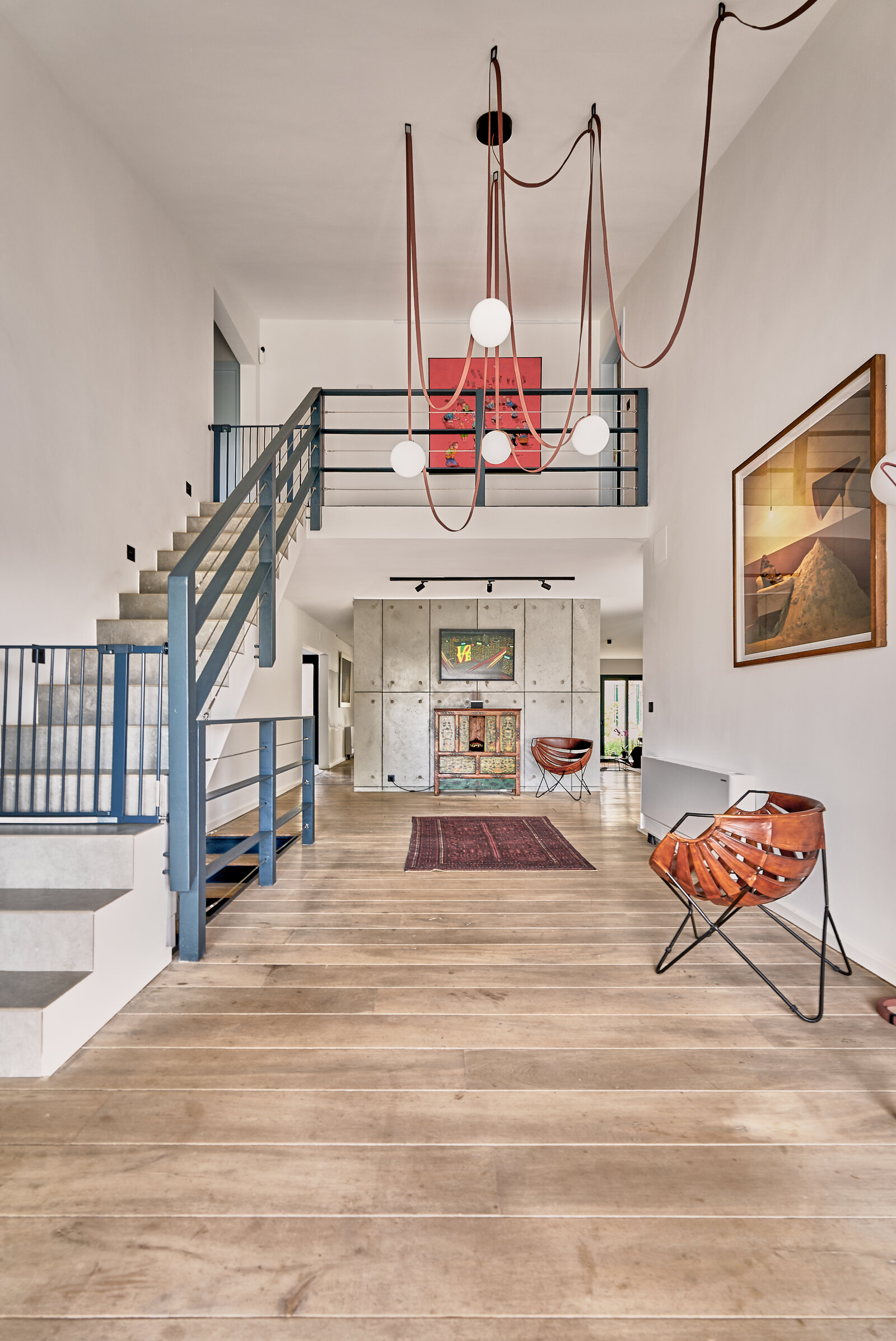Double-height hall with a steel staircase to a gallery, wide wooden floorboards, leather chairs and red pendant cables with glass globes.