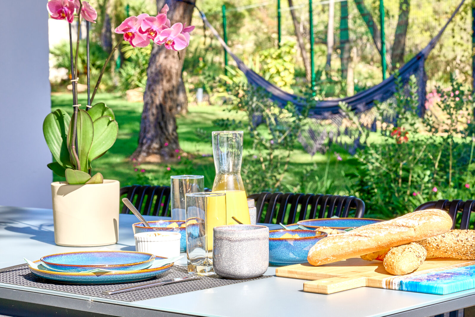 A breakfast table outside with a baguette, orange juice, blue bowls and an orchid, a hammock in the garden behind.