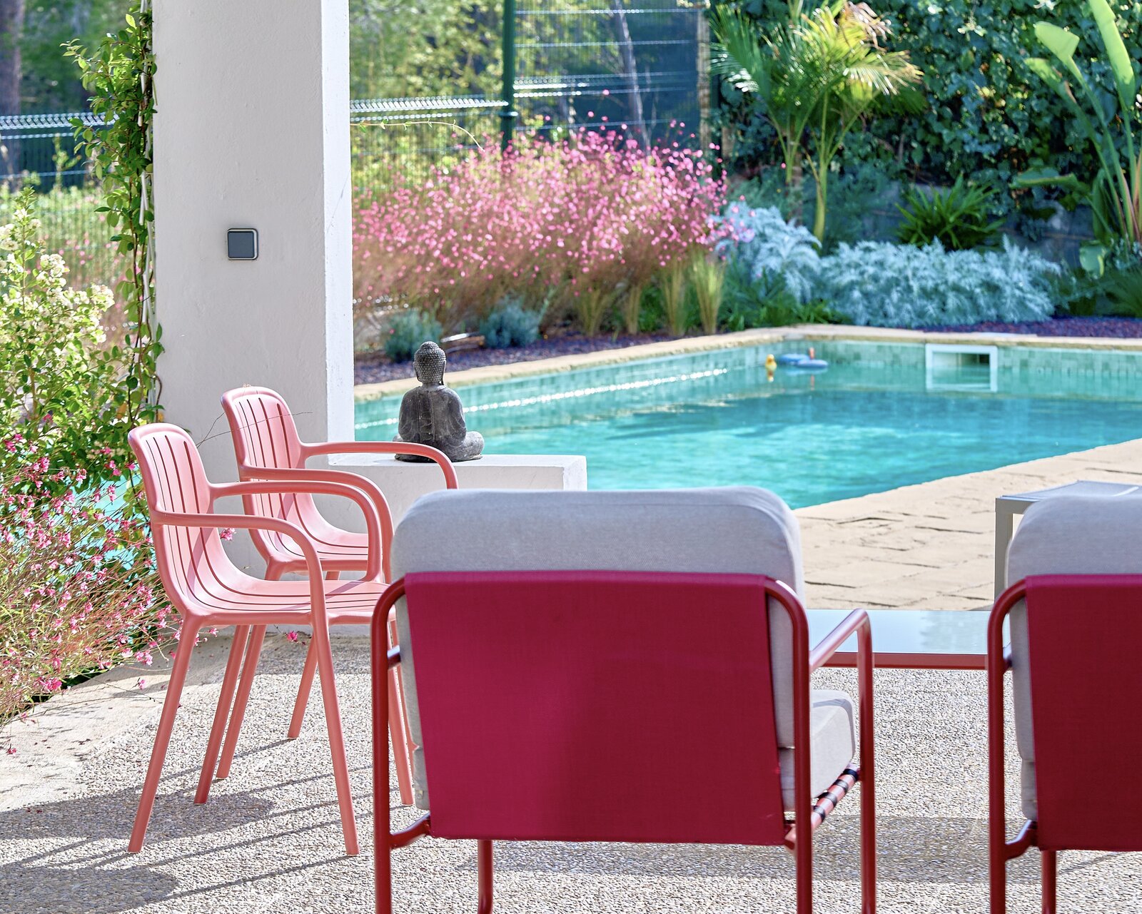 Pink chairs and red armchairs on a shaded terrace, with a small Buddha statue and the pool and flowering garden behind.