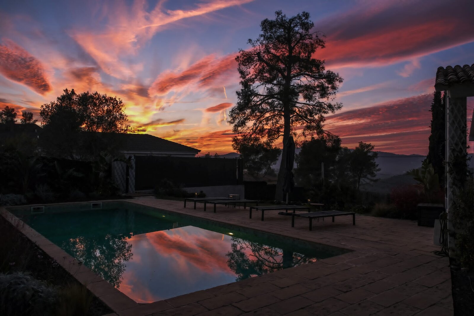 The pool at dusk reflecting a red sunset sky, with loungers on the paving and a tall pine in silhouette.