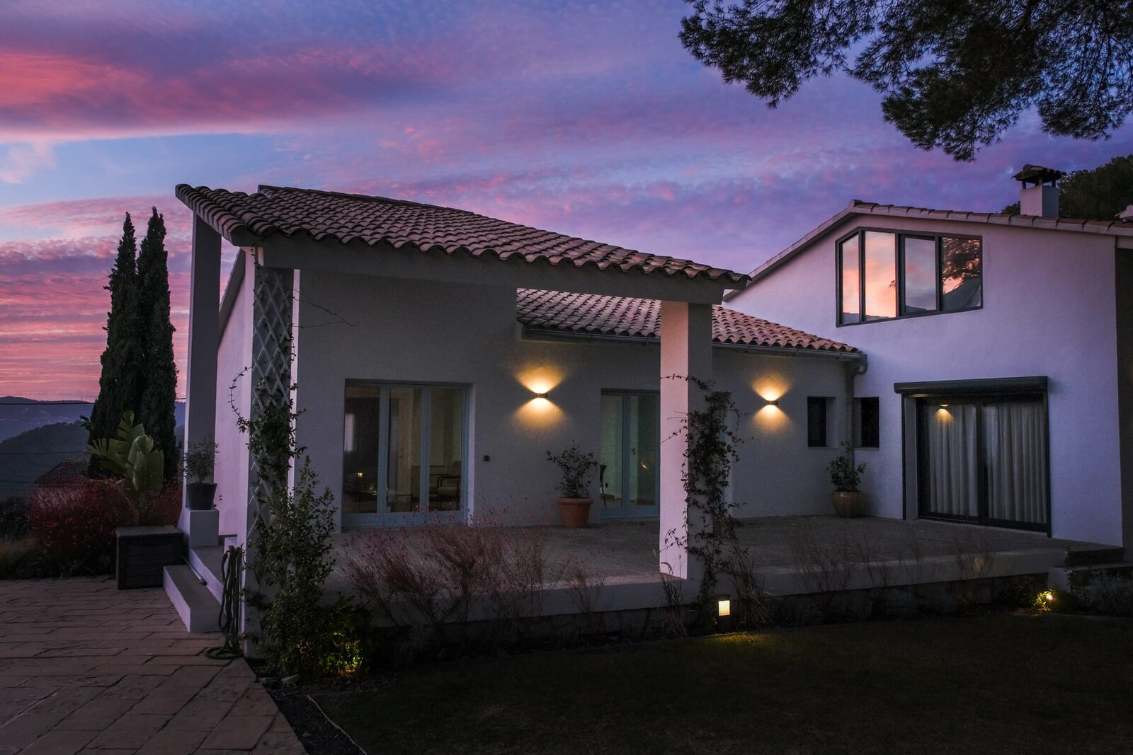 The white house lit at dusk under a pink and purple sky, with a covered terrace and wall lights.