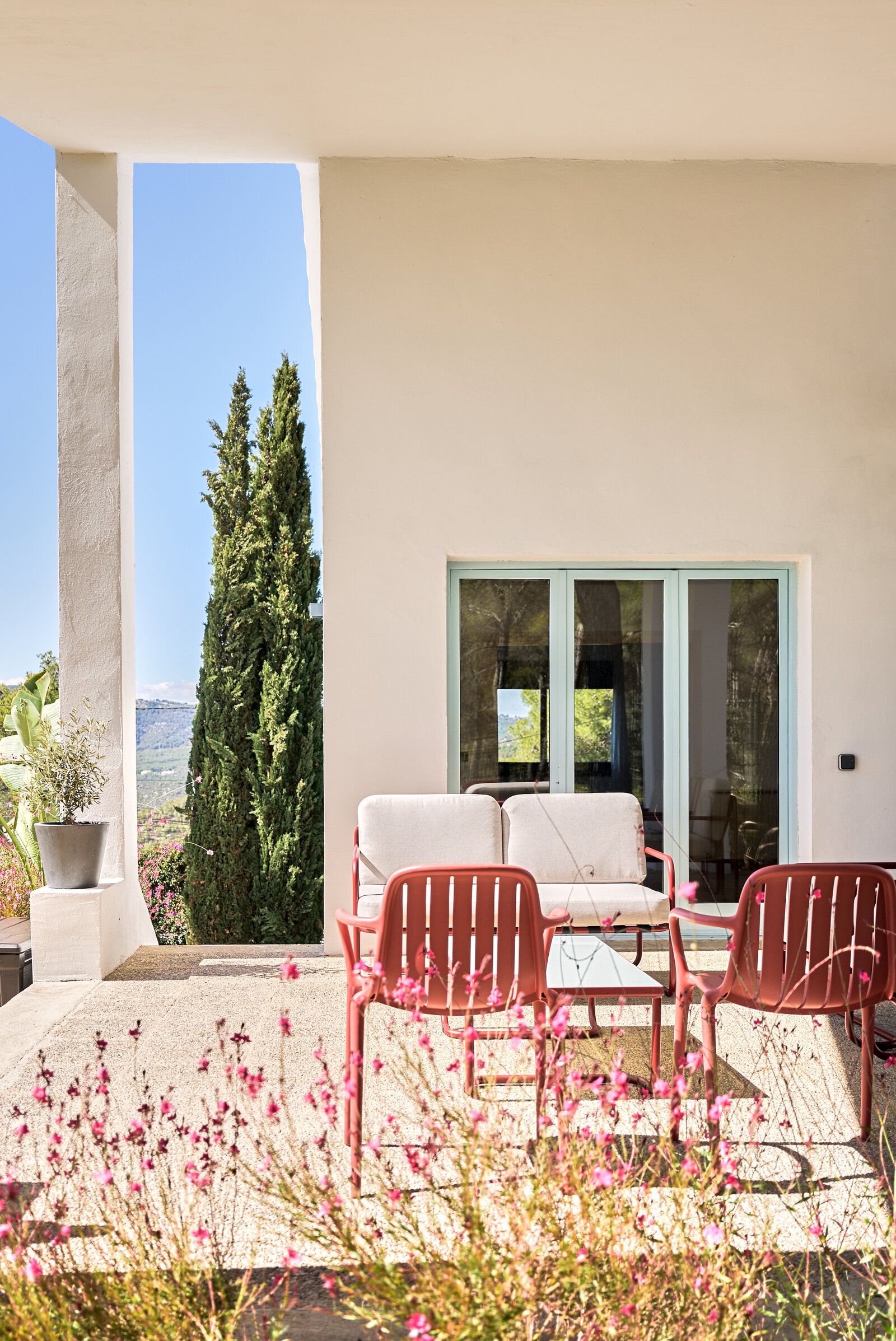 A covered terrace with a white sofa, red chairs and a glass table, a glazed door behind and pink flowers in front.
