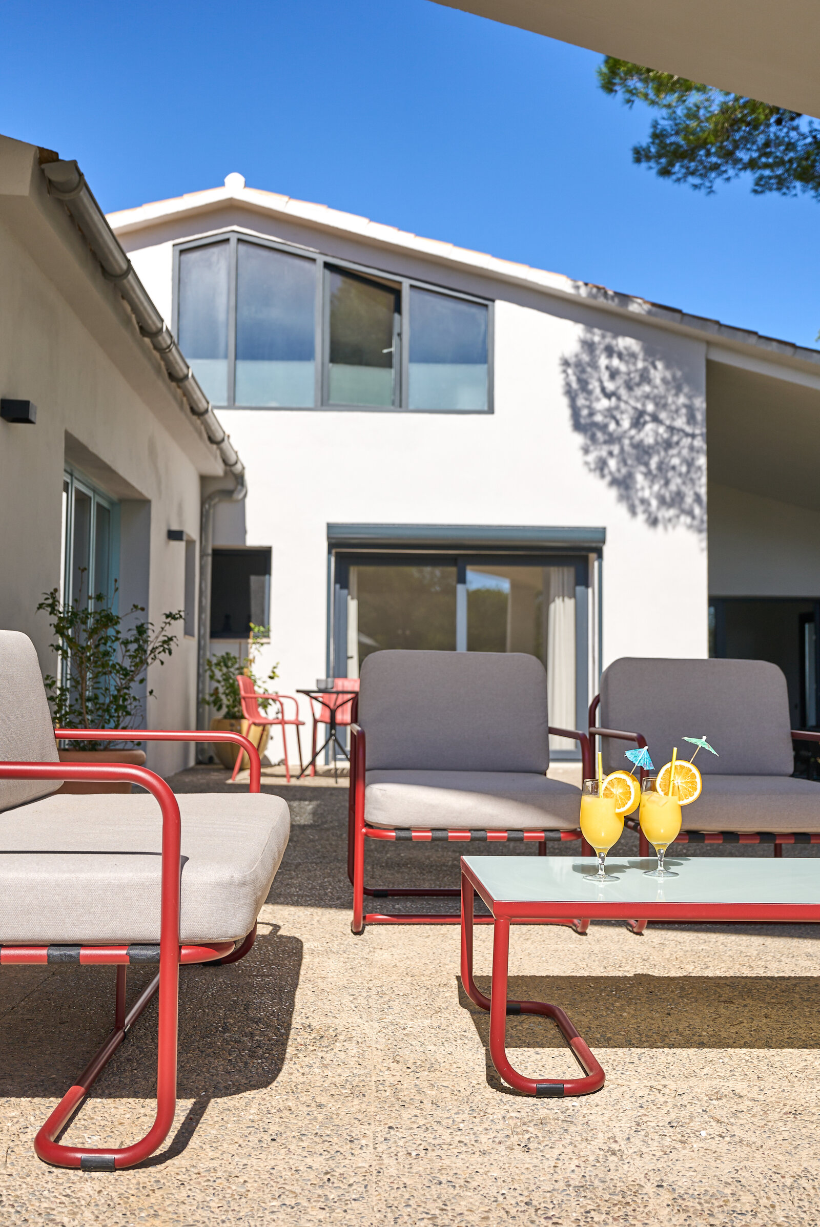 Grey and red lounge chairs around a low table with two cocktails in the courtyard, the white two-storey house behind.