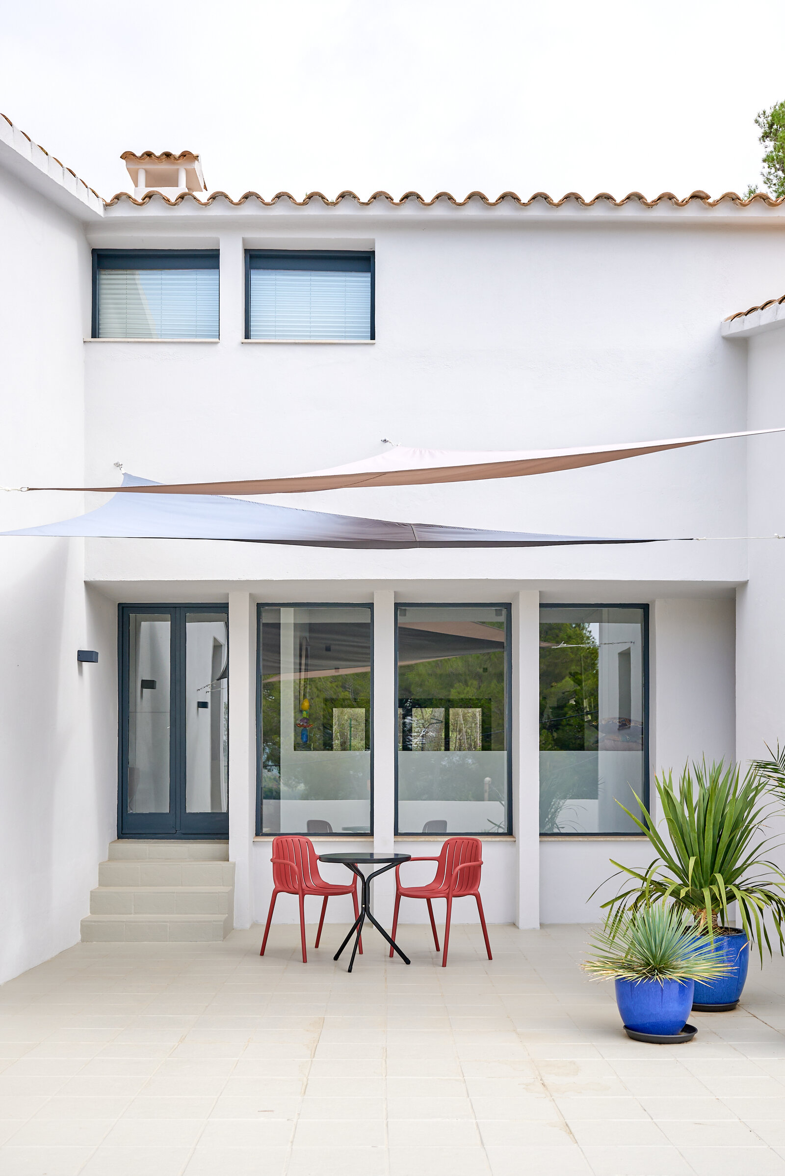 The white courtyard with shade sails overhead, a small red bistro set and potted plants in blue pots.