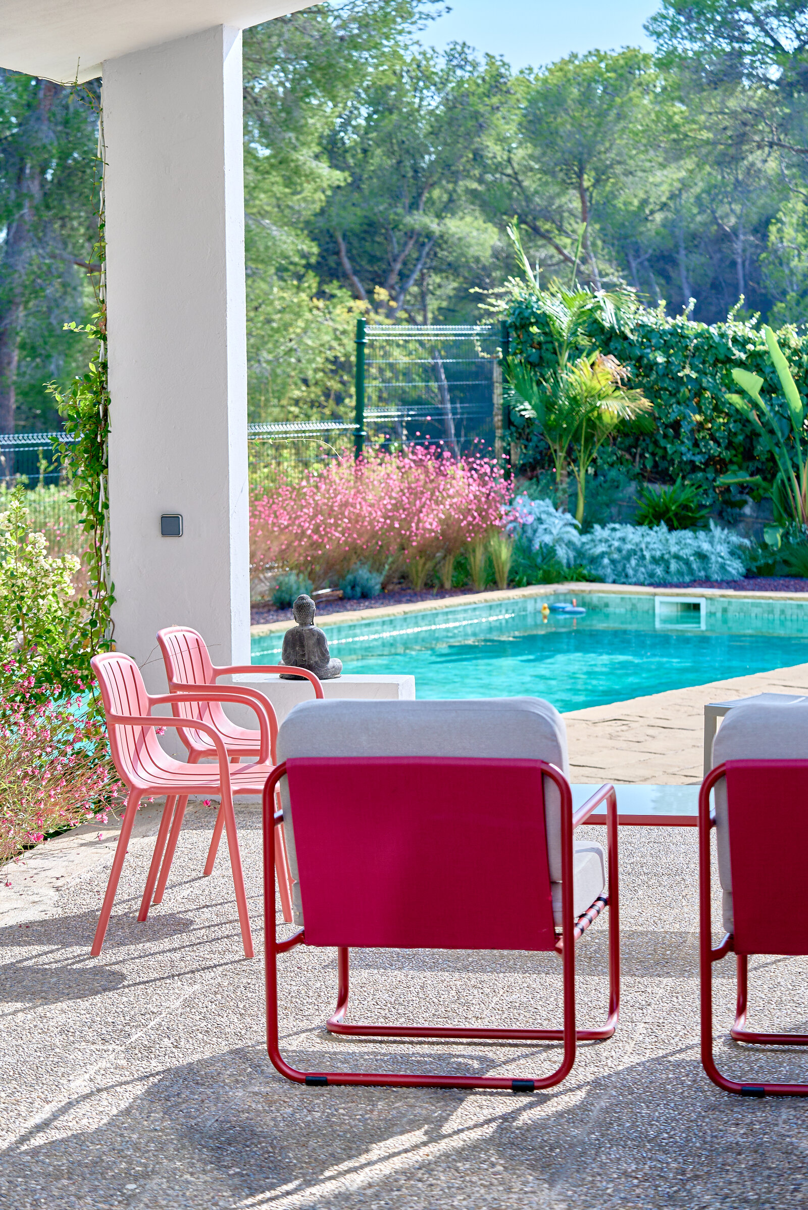 Red and pink chairs on the terrace facing the pool, with a small Buddha statue and pink flowers along the far edge.