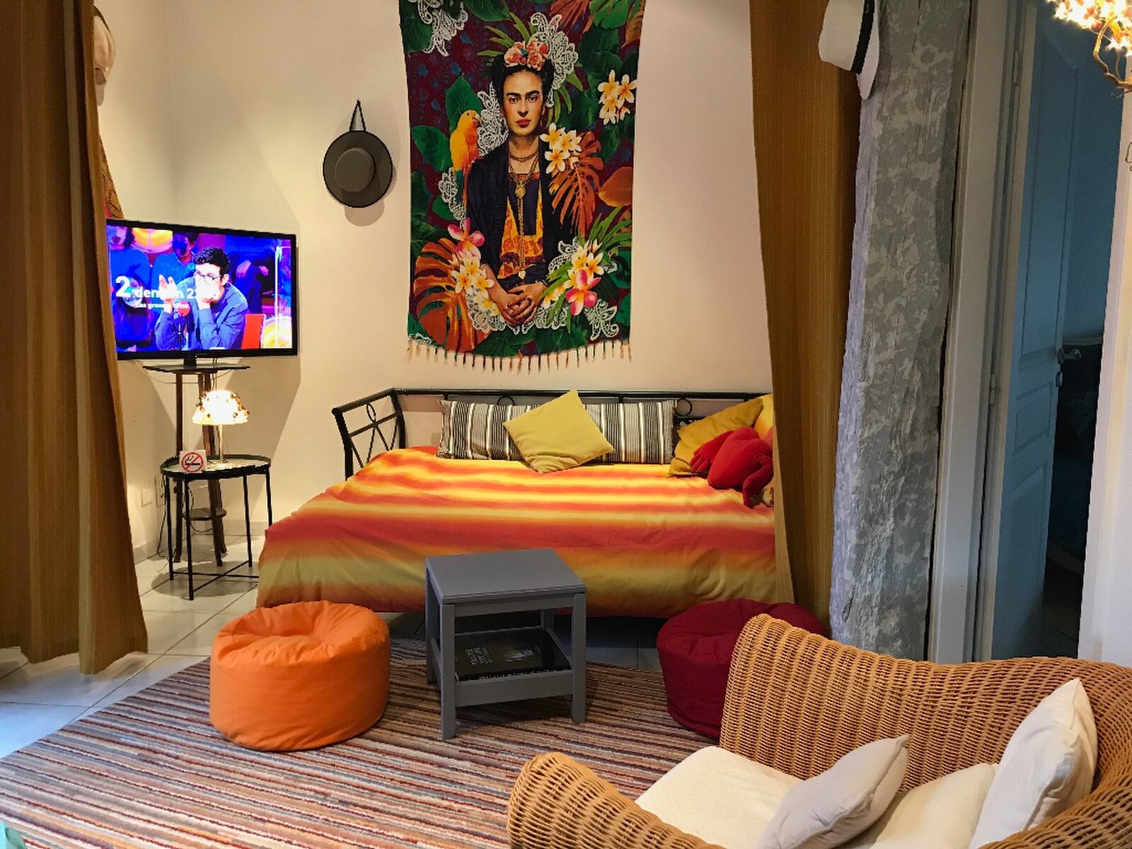 A day bed with striped orange and red covers under a Frida Kahlo wall hanging, with a television, an orange pouf and a wicker armchair in front.