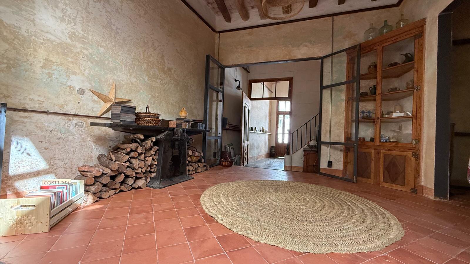 A large hall with a terracotta floor, a round jute rug, a stack of firewood under a workbench and a glazed cabinet with crockery.