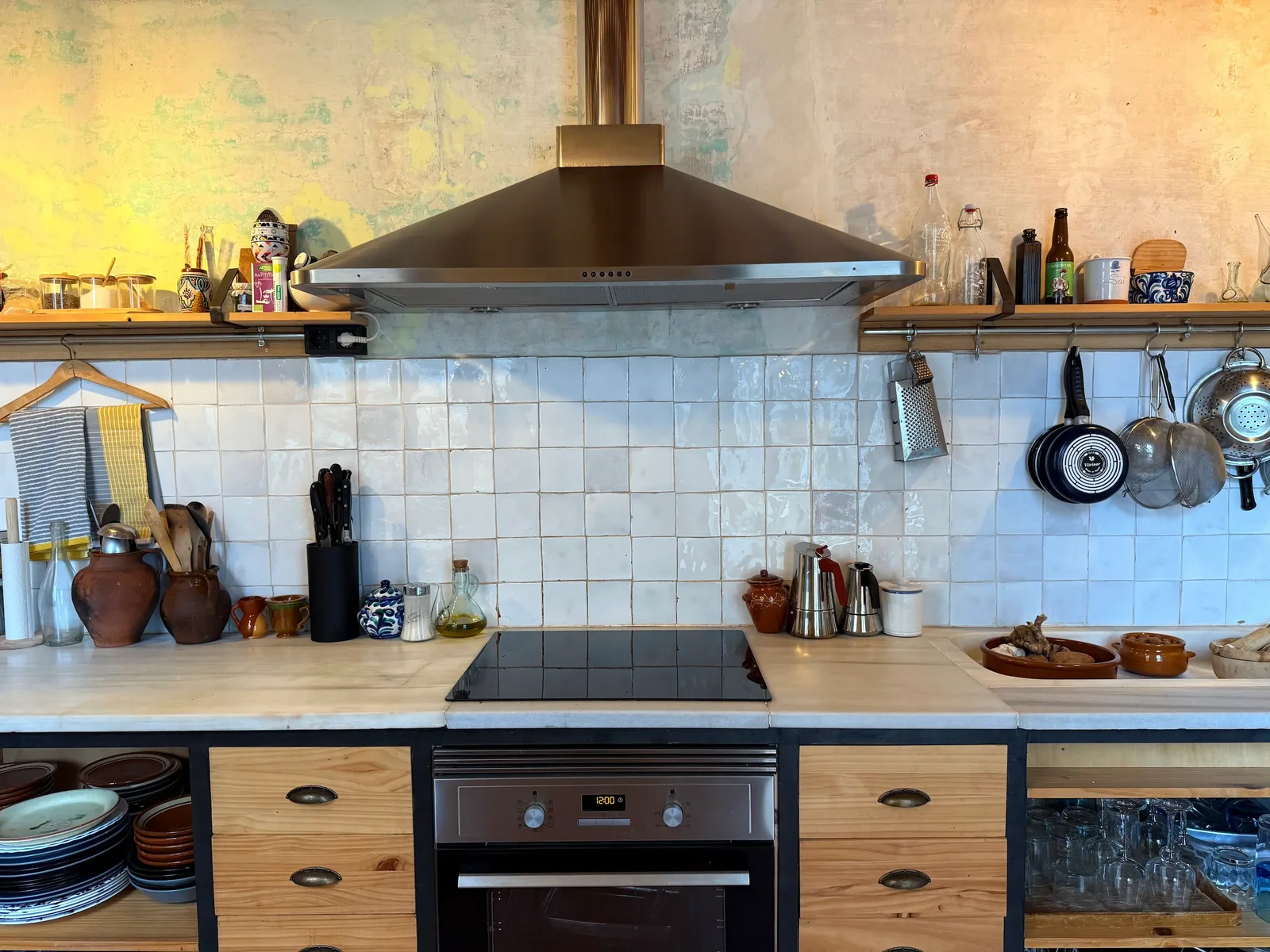 The kitchen with white tiles, a steel extractor hood over an induction hob, wooden drawers and shelves of pots and jars.