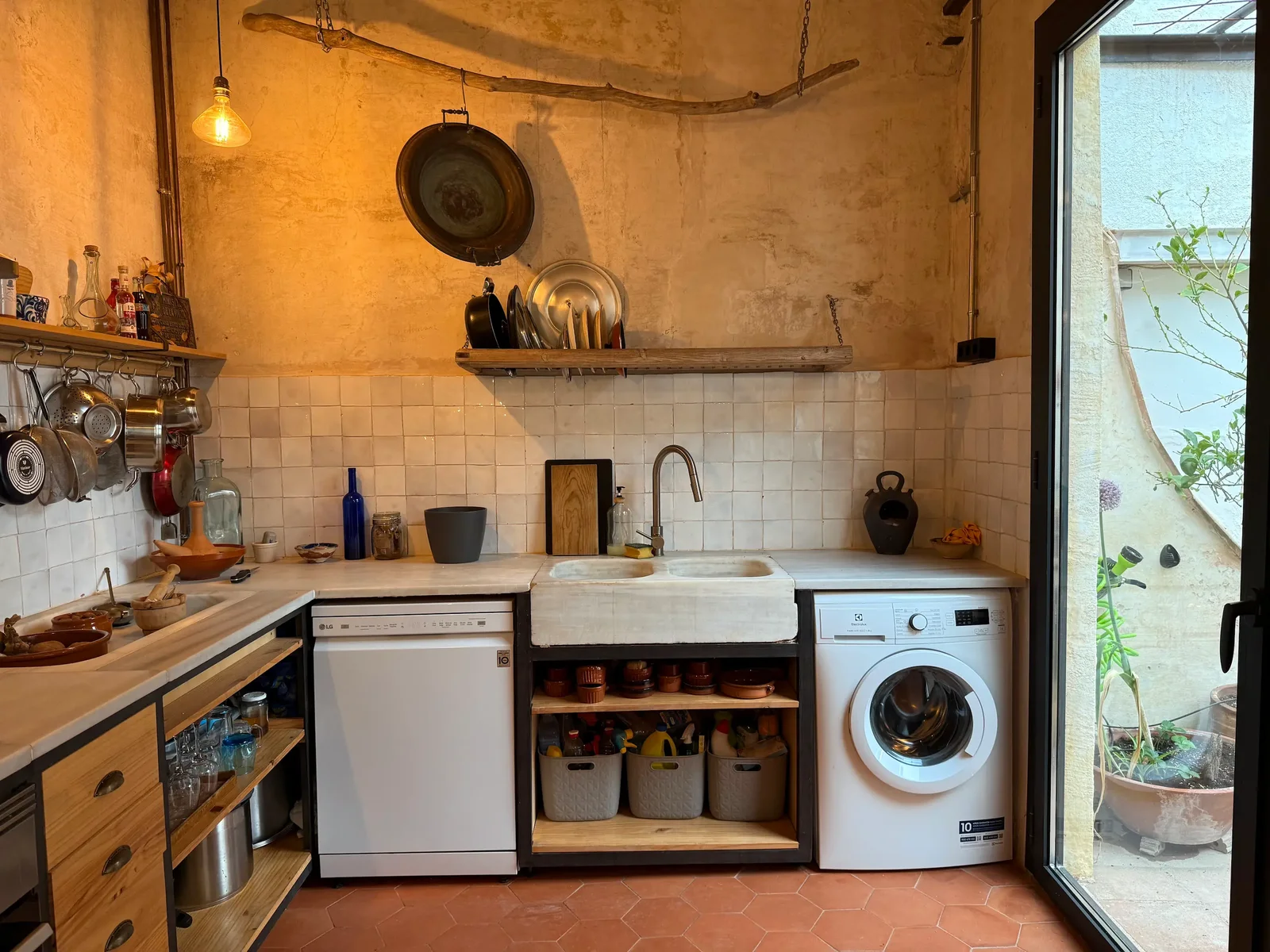 A rustic kitchen with a stone sink, a dishwasher and a washing machine on a terracotta floor, and a glass door to the courtyard.