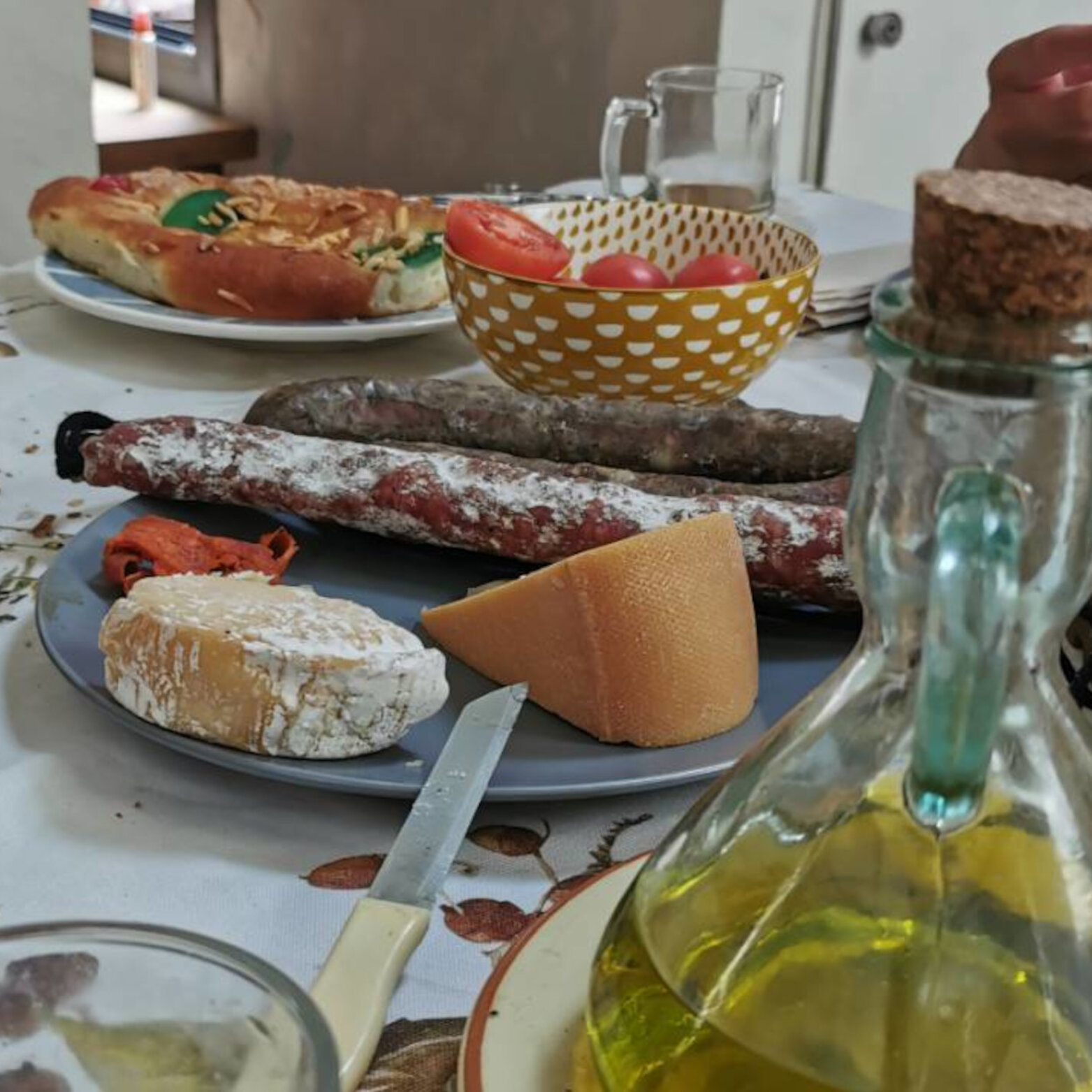 A table set with cured sausages, cheese, a bowl of tomatoes, a pastry and a glass jug of olive oil.