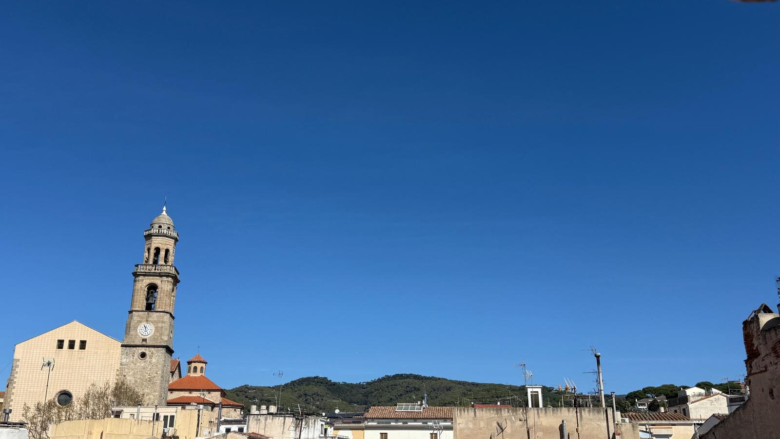 A view over the rooftops to a church bell tower and green hills under a clear blue sky.