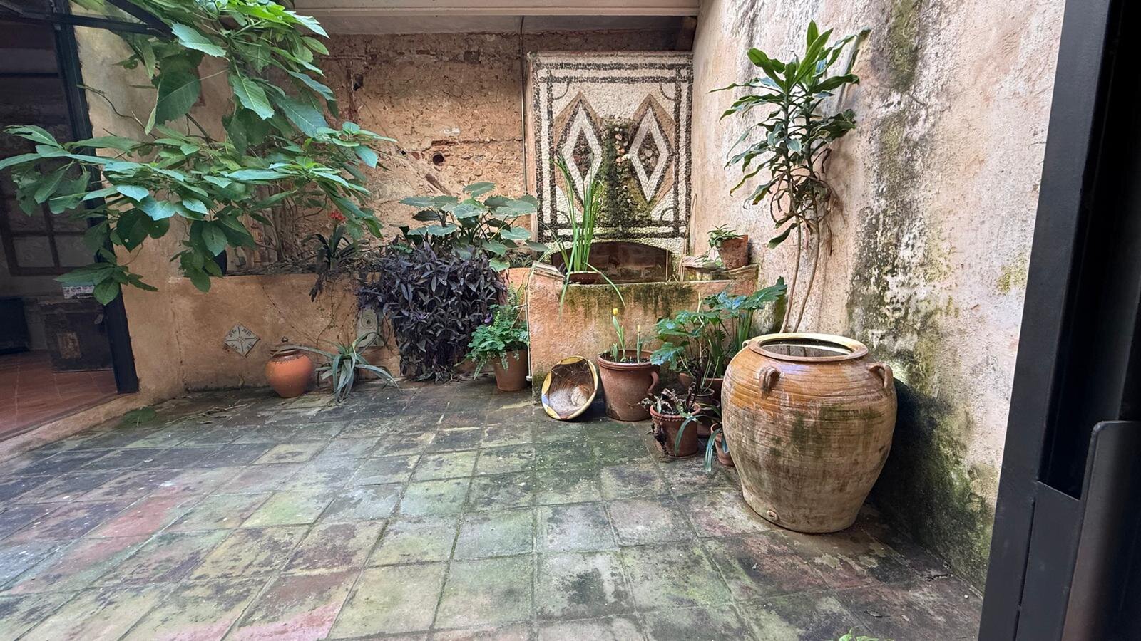 An inner courtyard with weathered floor tiles, large clay jars, potted plants and a mosaic panel on the wall.