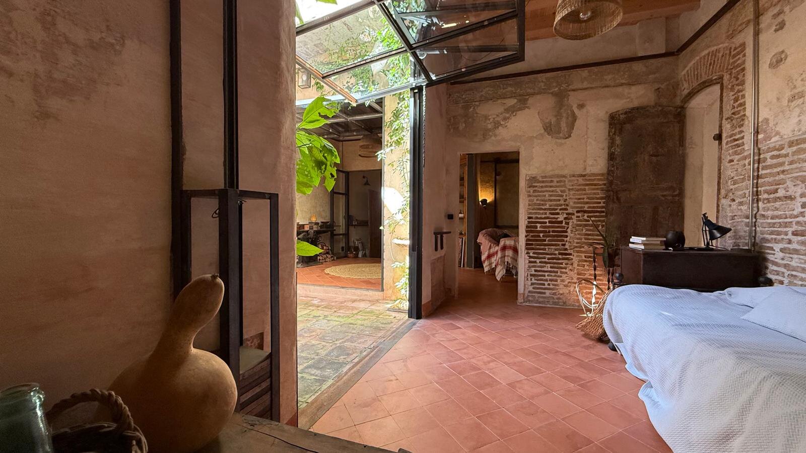 A bedroom with terracotta floor tiles and exposed brick, a glazed door open onto the courtyard and a second bed in the room beyond.