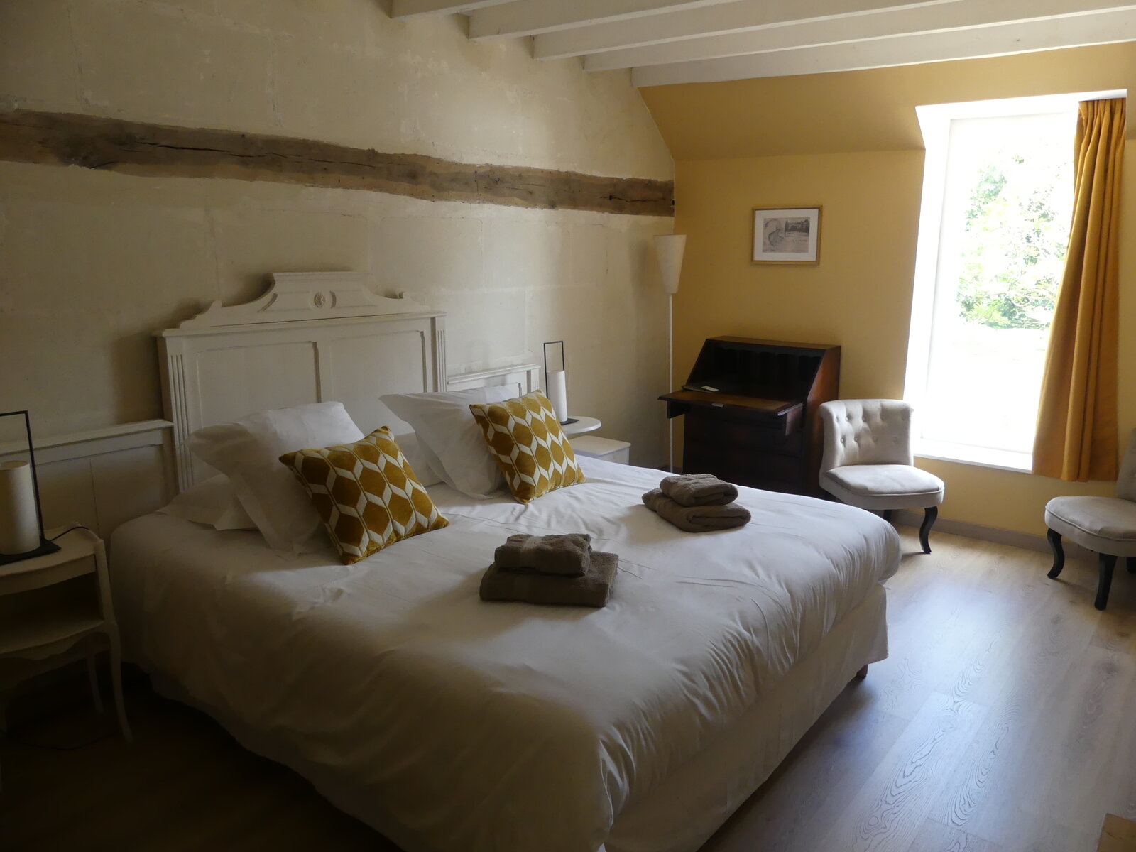 A guest room with a tuffeau stone wall, an exposed beam and a window onto the grounds.