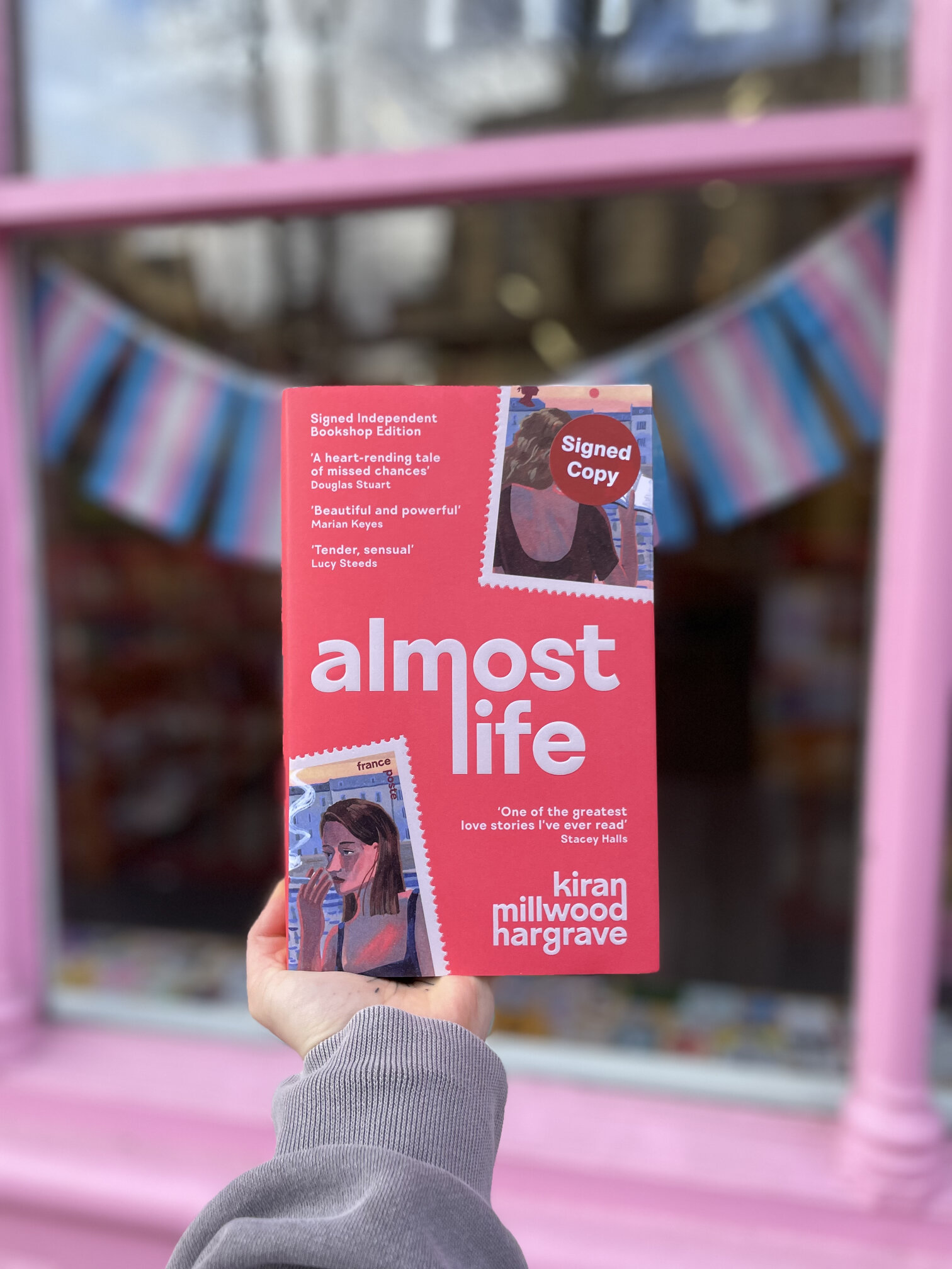 A signed copy of Almost Life by Kiran Millwood Hargrave, held up outside the pink shopfront.