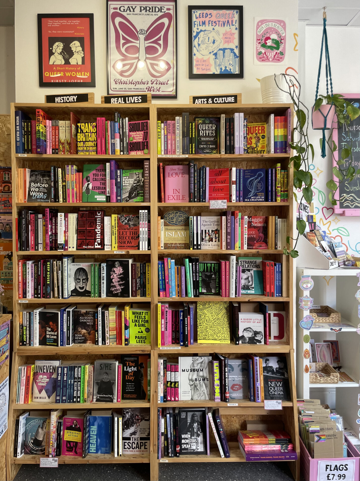 Shelves of queer non-fiction under posters for Gay Pride, Leeds Queer Film Festival and Christopher Street West, grouped as History, Real Lives and Arts & Culture.