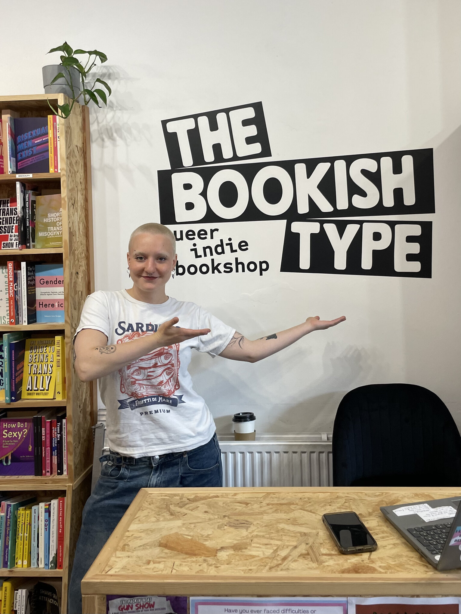 A member of staff points to the hand-painted "The Bookish Type, queer indie bookshop" mural above the till.