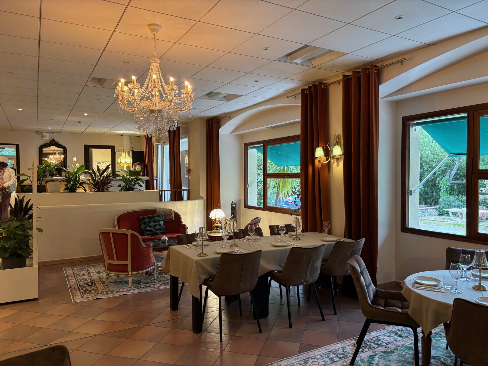 The restaurant L'Émeraude under a crystal chandelier, with a red salon corner by the window and tables laid for dinner.