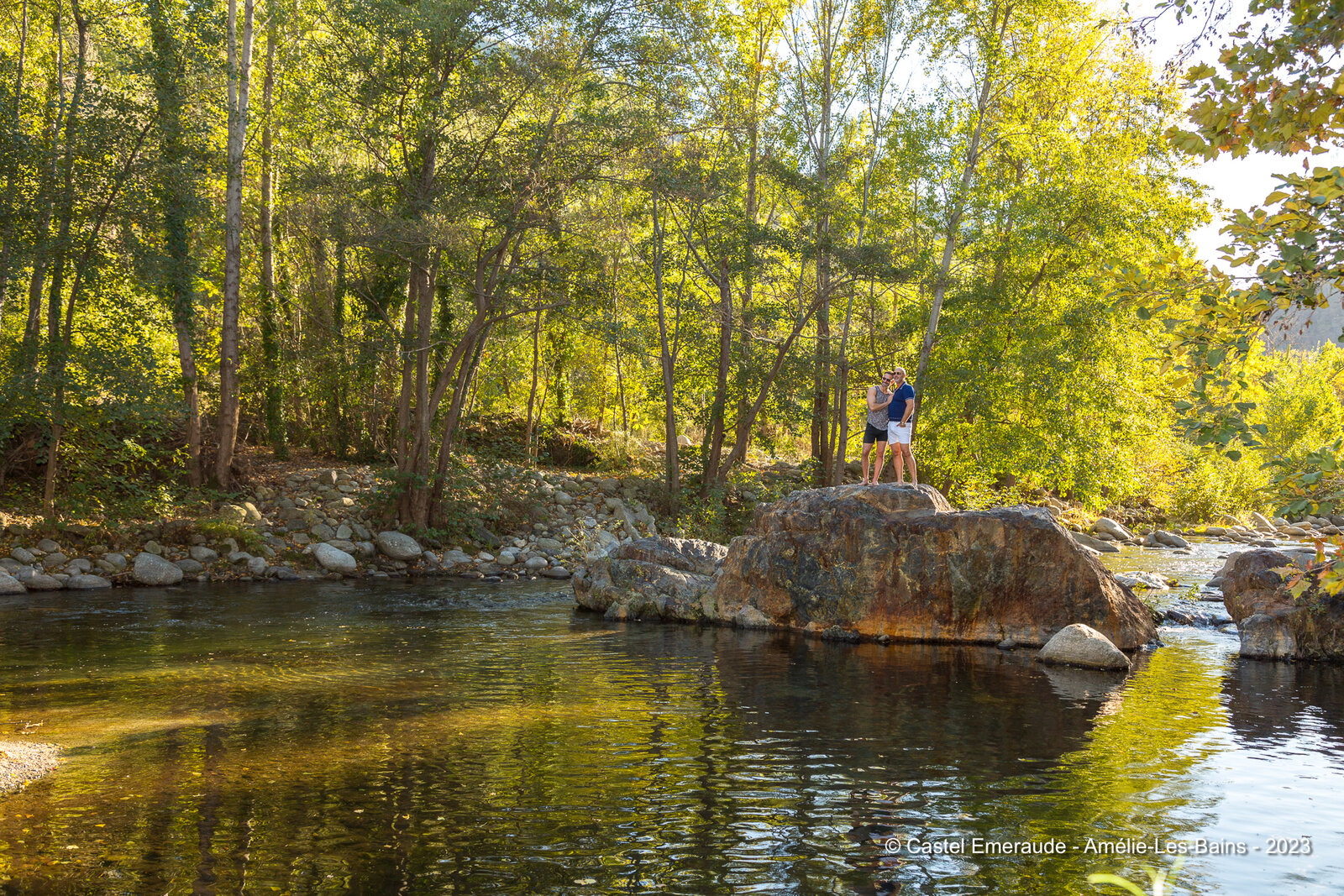 Two members of the family standing on a large boulder in the river, trees all along the bank.