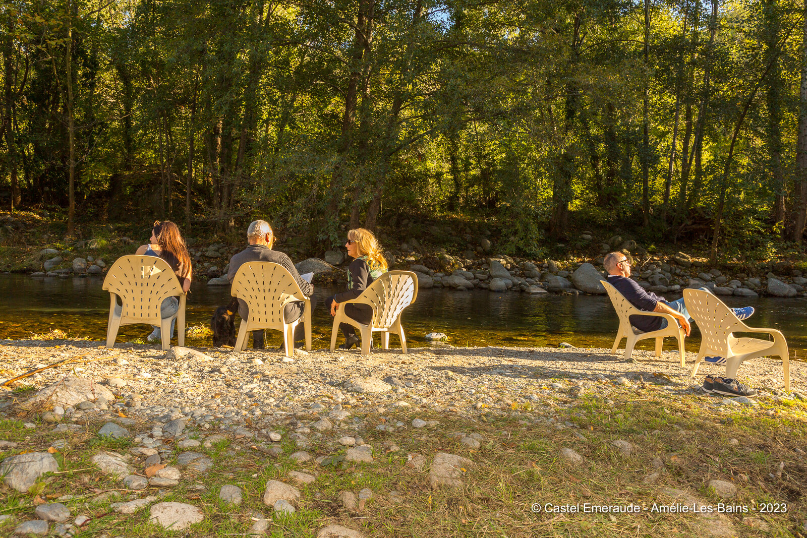 The family in garden chairs on the pebble bank, in the late afternoon sun by the river.