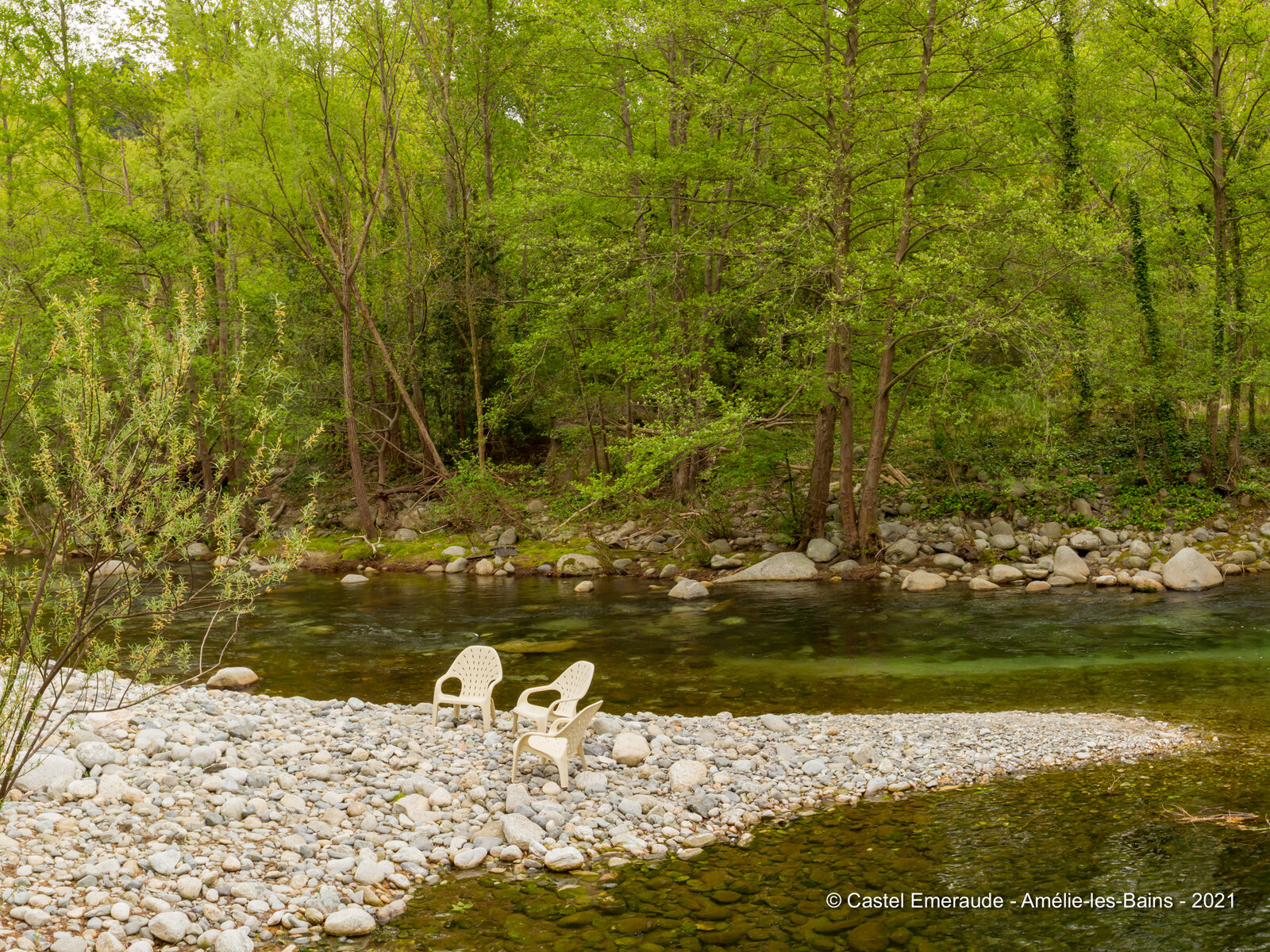 Three garden chairs on a pebble bank by the river, with the woods coming down to the water.