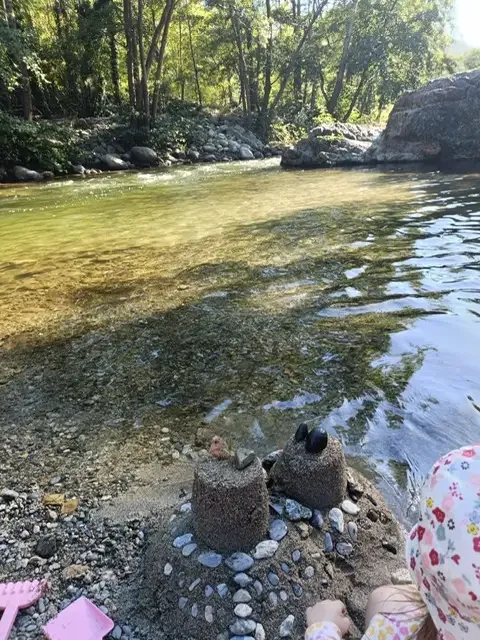 A sandcastle decorated with pebbles on the riverbank, the water clear over the stones.
