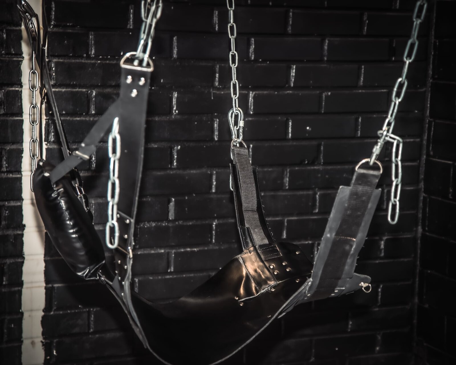 Leather sling hanging from chains in front of a black brick wall