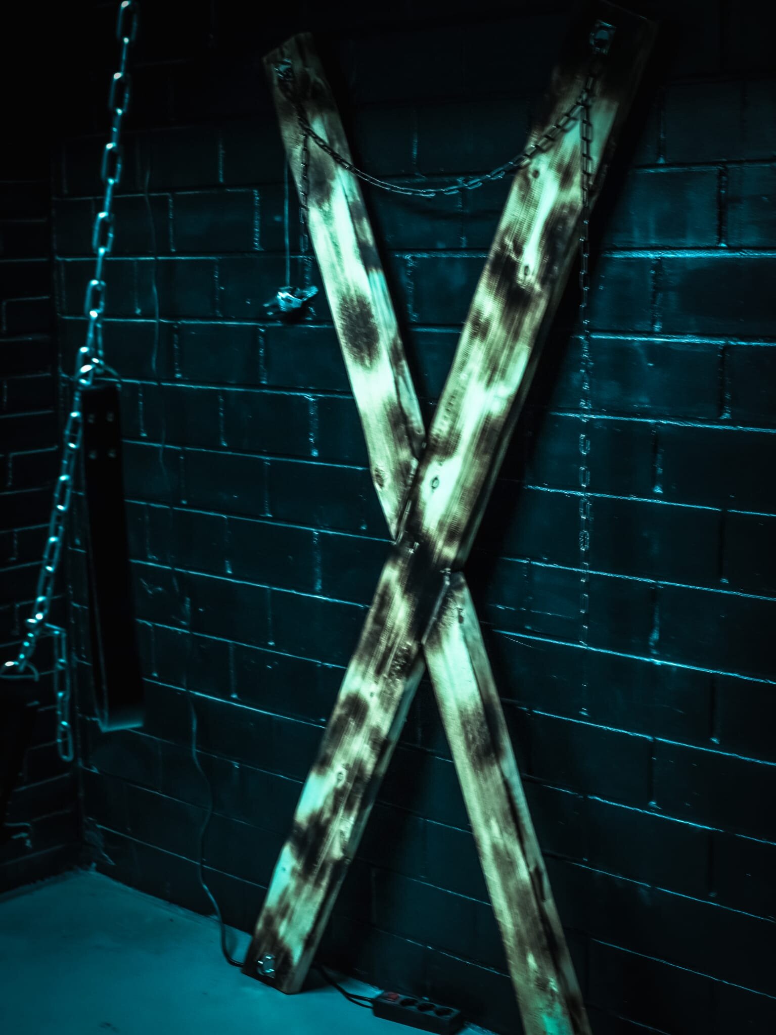 Wooden St Andrews cross leaning against a black brick wall, with chains and a leather strap beside it