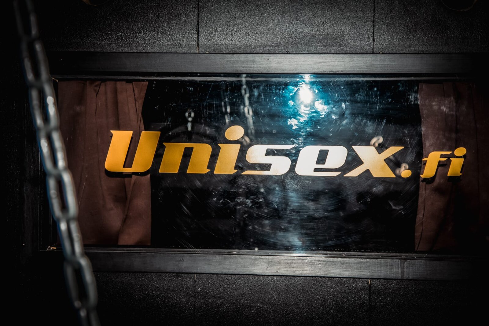 Unisex.fi sign in the dungeon area, with curtains behind a window and a chain in the foreground