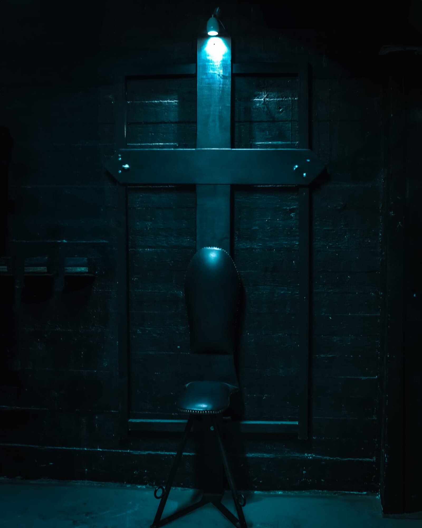 Dungeon room with a wall-mounted wooden cross and a leather-topped stool, lit in blue