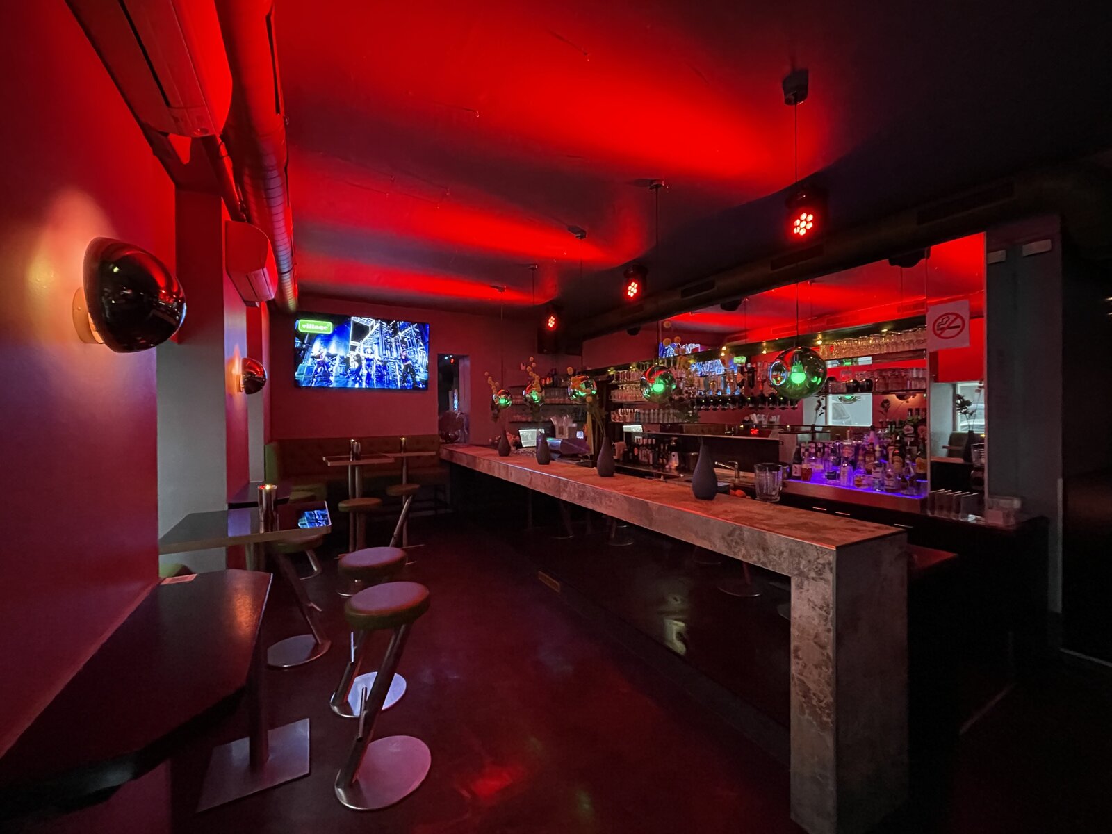 The full length of the room under red light, bar stools along the stone counter on the right and high tables under the screen on the left