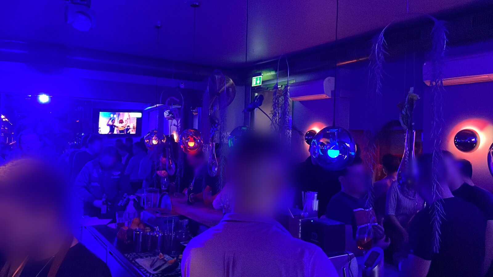 A busy evening at Village Bar, guests standing several deep along the counter under blue light and the chrome pendant lamps