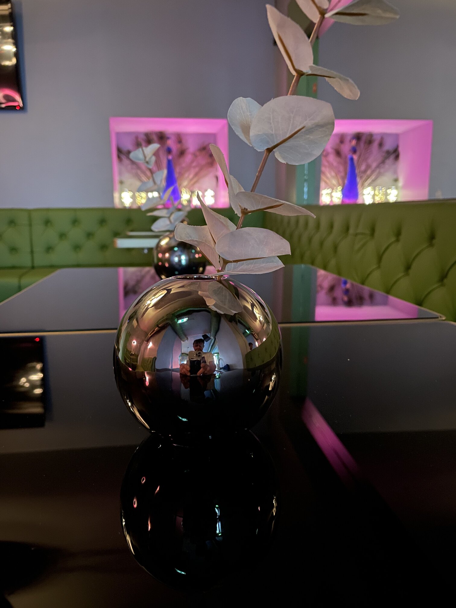 Dried eucalyptus in a chrome vase on a mirrored table, in front of the green tufted banquette and the pink-lit wall niches