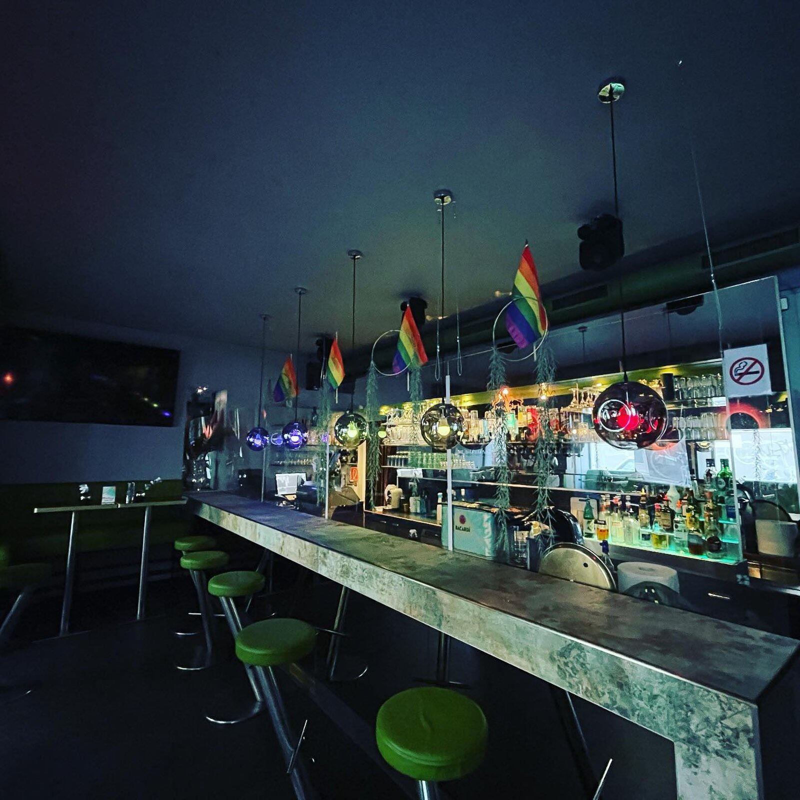 Rainbow flags in metal hoops over the counter, above the green bar stools and the lit spirits shelf