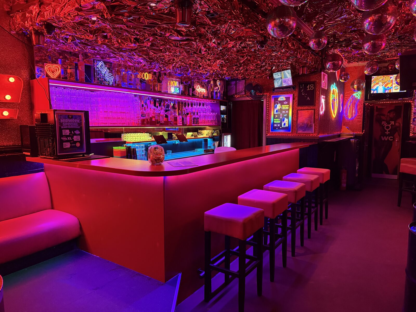 The bar counter under a ceiling of crumpled pink foil, with neon signs and disco balls above the stools