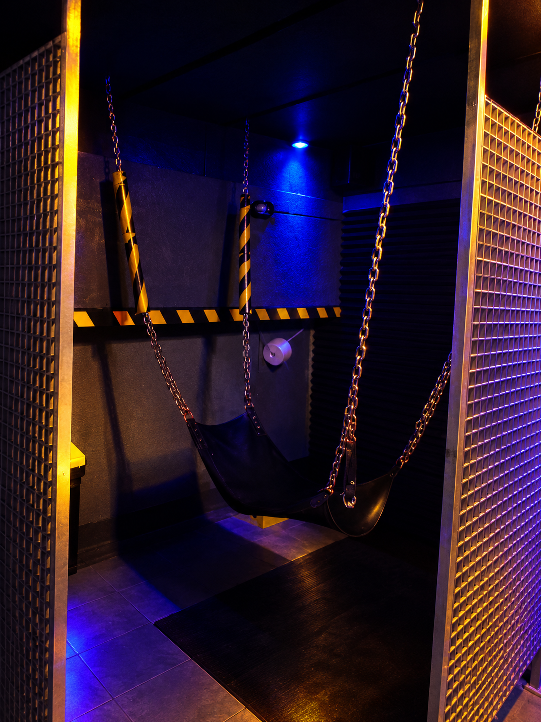 A leather sling on chains in a booth screened off by metal grating.